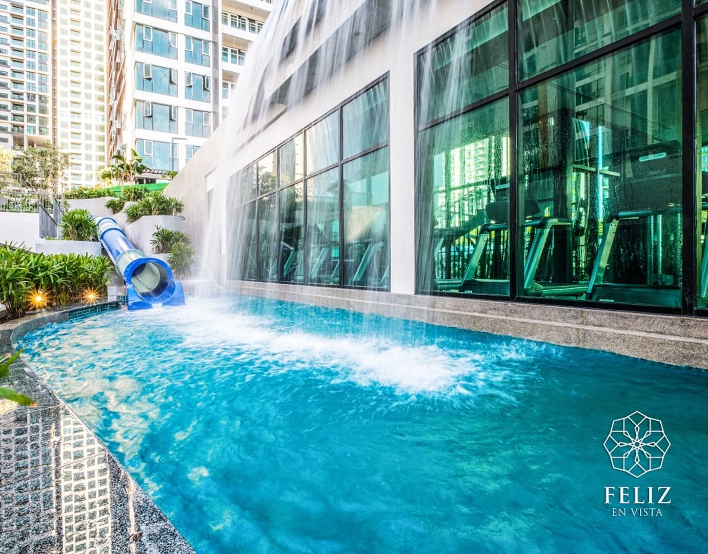 Khu vui chơi dưới nước tại Feliz En Vista Quận 2 có cầu trượt xanh, thác nước và hồ bơi dành cho trẻ nhỏ, bên cạnh không gian phòng tập nhìn qua những ô cửa kính lớn.